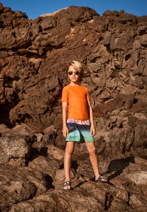 Rash guard orange vif à manches courtes, short de bain multicolore avec motif dégradé, et sandales sombres. Fond de terrain rocailleux.