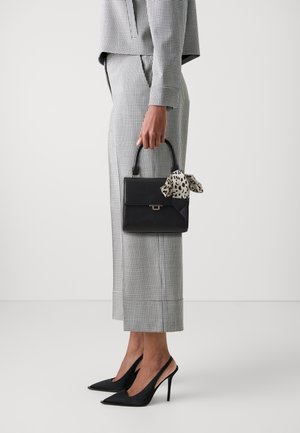 Frau in einem schwarz-weiß karierten Anzug, die eine schwarze Handtasche mit einem gemusterten Tuch am Henkel hält und schwarze Slingback-High-Heels trägt.