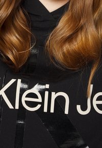 Czarna bluza z kapturem z wyraźnym białym logo "Klein Jeans". Posiada błyszczące czarne akcenty taśmy na przedniej stronie, tworząc teksturowany kontrast.
