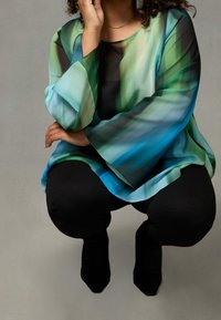 Blusa trasparente a maniche lunghe con un motivo a gradiente blu e verde. Abbinata a leggings neri e scarpe nere appuntite. Vista dettagliata del modello.