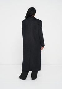 Cappotto nero lungo realizzato in un materiale testurizzato, caratterizzato da una silhouette dritta, ampi rever e che arriva fino a metà polpaccio.