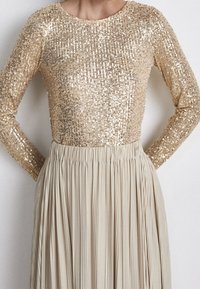 Gouden pailletten lange mouw top gecombineerd met een geplooide beige rok. De gladde textuur van de top staat in contrast met de luchtige, gestructureerde rok.