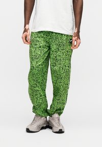 Zöld foltmintás melegítőnadrág rugalmas derékpánttal és oldalzsebekkel, szürke sportcipőkkel párosítva. Fekete márkajelzés látható.