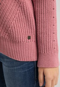 Pletený svetr v jemně růžové barvě s žebrovanými manžetami a lemem. Má strukturovaný vzor a malou hnědou tkaninovou značku blízko lemu.