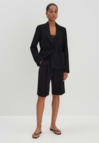 Blazer nero sartoriale con vita cinte e chiusura a singolo bottone, abbinato a pantaloni corti fino al ginocchio, caratterizzato da linee pulite e design minimalista.