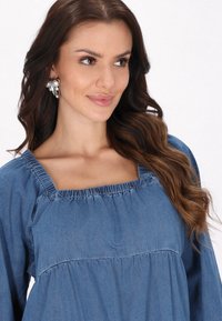 Blå denimtopp med fyrkantig halsringning, samlad detalj vid axlarna och bysten. Modellen har långt vågigt hår och bär stora pärlörhängen.