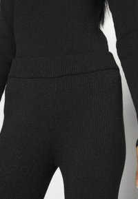 Haut noir en maille côtelée ajusté à manches longues et pantalon taille haute assorti sur une personne sur fond blanc.
