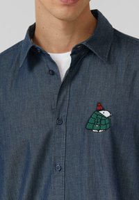 Granatowa koszula dżinsowa z zapinaniem na guziki, z haftowanym postacią z kreskówki w zielonym płaszczu i czerwonym kapeluszu na lewej piersi.