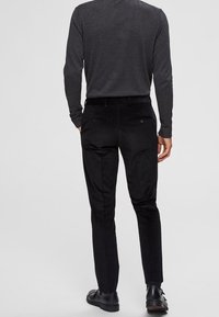 Svarta, åtsittande byxor med en slät yta och två bakfickor, i kombination med en mörk långärmad skjorta och svarta skor.