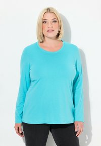 Türkise Langarm-T-Shirt aus weichem Stoff, mit rundem Ausschnitt und lockerer Passform, kombiniert mit dunklen Jeans.