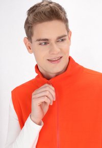 Oranžová fleecová vesta na zip s vysokým límcem, s hladkým povrchem a plným zipem, nošená přes bílou košili s dlouhým rukávem.