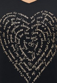 Μαύρη μπλούζα V με σχέδιο που παρουσιάζει στροβιλιζόμενο, καλλιγραφικό κείμενο σε μπεζ για την αγάπη και τη σύνδεση, συνοδευόμενο από δύο καρδιές.