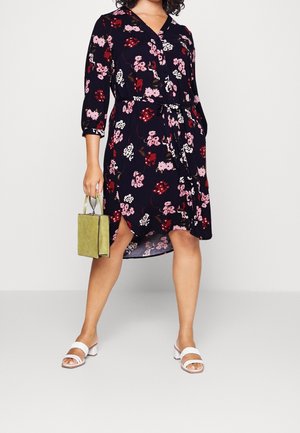 Frau trägt ein dunkles, knielanges Kleid mit Blumenmuster und 3/4-Ärmeln, hält eine kleine grüne Handtasche und trägt weiße Sandaletten mit Absatz.