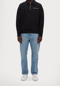 Svart zip-up sweatshirt för män med krage, framficka och vit logotyp. Matchad med ljusblå jeans och vita sneakers.