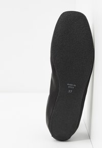 Semelle de chaussure plate noire avec grip texturé, indiquant la taille 37 et le texte "Fabriqué en Espagne", sur fond blanc avec un coin de mur.