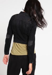 Chaqueta corta de mezclilla negra con detalles desgastados, puños abotonados y cuello estándar, llevada sobre una camiseta de color verde oliva claro.