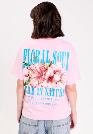 Persoon met schouderlang bruin haar, een roze T-shirt dragend met een groot bloemmotief en de tekst "FLORAL SOUL CALM IN NATURE" op de achterkant.