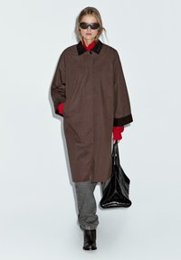 Cappotto marrone con colletto, polsini neri a contrasto e tasche laterali. Abbinato a jeans grigi e una borsa nera, indossato con occhiali da sole.