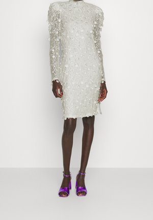 Robe blanche en dentelle à manches longues avec des pois texturés portée avec des talons ouverts violets brillants à bride cheville sur un mannequin à la peau foncée.