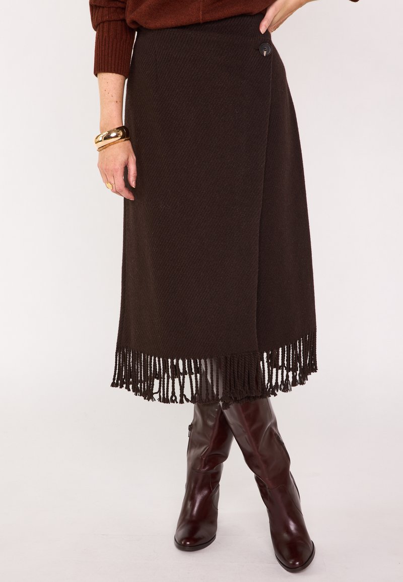 Gonna intrecciata marrone con orlo a frange, dotata di chiusura con bottone laterale. Abbinata a stivali marroni al ginocchio per un look coordinato.