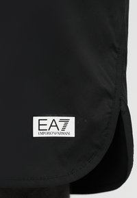 Μαύρο ύφασμα με κυρτή ραφή και ορθογώνια λευκή ετικέτα που δείχνει το λογότυπο "EA7 Emporio Armani" ραμμένο κοντά στην άκρη.