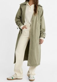 Olivenvihreä puuvillatrenssi takki, jossa on kaksiriviset napit, säädettävät hihansuut ja sivuaukot, päällä kermaa värinen villapaita ja vaaleat housut.