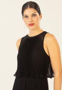 Schwarzes plissee Kleid mit einem halbtransparenten Überzug, ärmellosem Design und rundem Ausschnitt. Es zeichnet sich durch eine subtile Textur und eine fließende Silhouette aus.