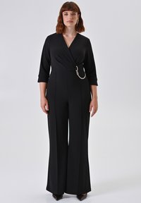 Tuta nera con maniche a tre quarti, design avvolgente e una grande fibbia metallica con dettaglio a catena in vita; pantaloni a gamba larga.