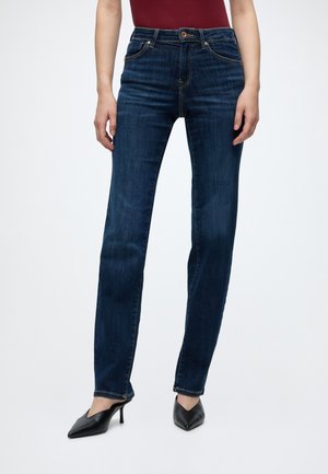 Donna che indossa jeans blu scuro a gamba dritta, tacchi alti neri a punta e una maglietta rossa con le mani rilassate ai lati.