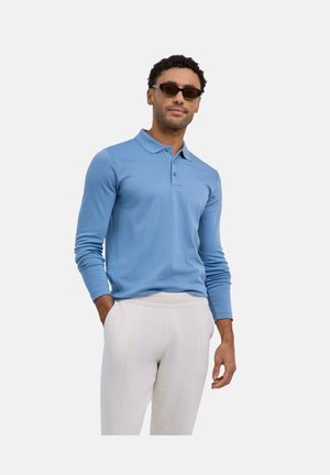 Uomo che indossa una polo a maniche lunghe blu, pantaloni bianchi e occhiali da sole scuri, in piedi con una mano in tasca su sfondo bianco.