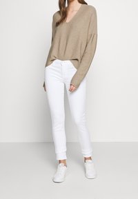 Beżowy sweter v-neck o luźnym fasonie, w zestawie z białymi dżinsami o slim fit. Białe tenisówki dopełniają stylizację. Gładka faktura, minimalistyczny design.