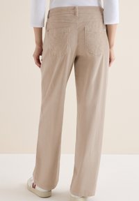 Beżowe spodnie o szerokich nogawkach z dwiema tylnymi kieszeniami z przeszyciami i gładką powierzchnią, w zestawieniu z białymi i czerwonymi sneakersami.