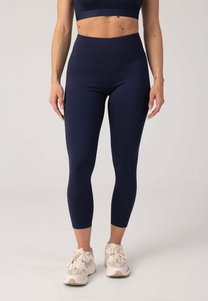 Kobieta ubrana w granatowe krótkie legginsy z wysokim stanem oraz dopasowany sportowy biustonosz, białe adidasy, stoi na tle jednolitego jasnego tła.