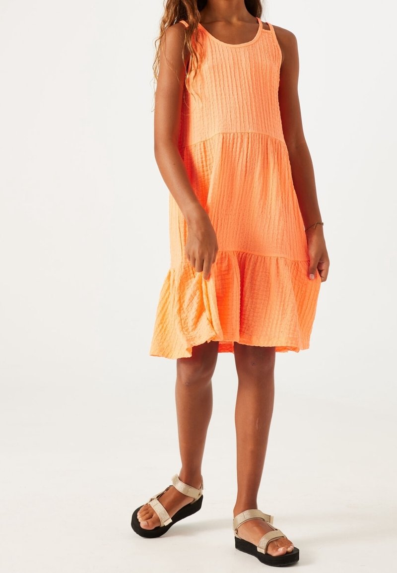 Vestido sin mangas de color naranja brillante con tejido texturizado, diseño en capas y escote redondeado, combinado con sandalias simples en negro y plata.