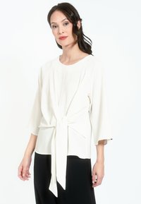Blusa blanca con escote redondo, mangas de tres cuartos y detalle de nudo atado en la cintura, hecha de una tela suave y ligera.