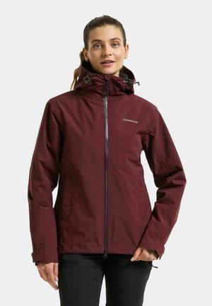 Frau mit einer bordeauxfarbenen wasserdichten Jacke mit Kapuze und schwarzer Hose steht vor einem schlichten hellen Hintergrund.
