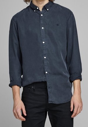 Homme portant une chemise bleu foncé boutonnée avec les manches retroussées et un pantalon noir sur un fond gris uni.