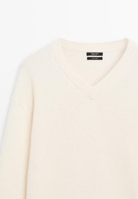 Cremefarbener Strickpullover mit V-Ausschnitt, geripptem Kragen und lockerer Passform. Hergestellt aus weichem Material und mit minimalen Nähdetails.
