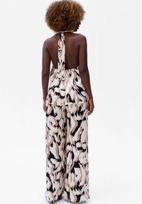 Vue arrière d'une combinaison sans manches en tissu imprimé avec un col halter et un dos dégagé, présentant des motifs abstraits beiges, noirs et marron.