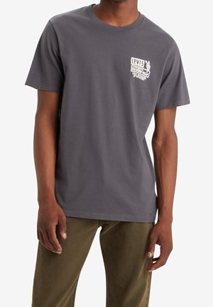 Mężczyzna noszący ciemnoszarą koszulkę Levi’s z białym logo Western Wear na piersi, w połączeniu z oliwkowozielonymi spodniami.