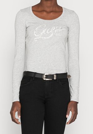 Person, die ein hellgraues Langarmshirt mit dem Text "Guess 1981" und schwarze Hose mit schwarzem Gürtel mit silberner Schnalle trägt.
