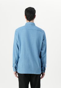 Camicia in denim azzurro chiaro con maniche lunghe, polsini a bottone, colletto e dettagli cuciti, caratterizzata da un orlo leggermente arcuato sul retro.