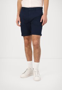 Pantalones cortos azul marino de corte recto, con cierre de botón y dobladillo enrollado. Llevados con zapatillas blancas y calcetines de longitud media. Material de algodón.