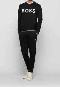 Hombre con sudadera y pantalones negros con el texto "BOSS", combinados con zapatillas blancas, de pie frente a un fondo gris liso.