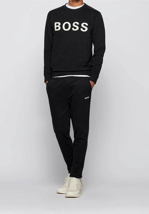 Man in een zwart sweatshirt en broek met de tekst "BOSS", gecombineerd met witte sneakers, staande tegen een effen grijze achtergrond.