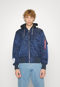 Mornarska bombarder jakna napravljena od satenskog materijala, s kapuljačom, zip zatvaranjem, rebrastim patentima i rubom, te crvenom oznakom na rukavu.