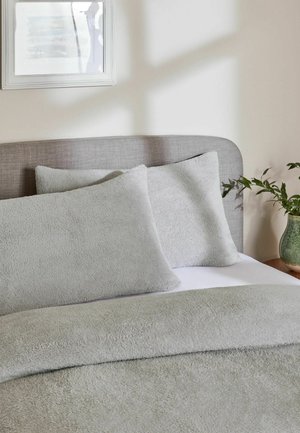 Bett mit einem grauen Stoffkopfteil, hellgrauen, strukturierten Kissen, einer passenden Decke, einem Wandbild darüber und einer grünen Pflanze in einer Vase auf einem hölzernen Nachttisch.