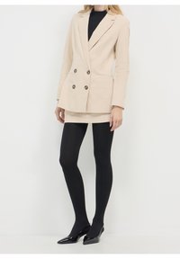 Beżowa, dwurzędowa marynarka z sztruksu z ciemnymi guzikami, zestawiona z dopasowaną beżową spódnicą, czarnymi rajstopami i czarnymi szpilkami z zaostrzonym noskiem.