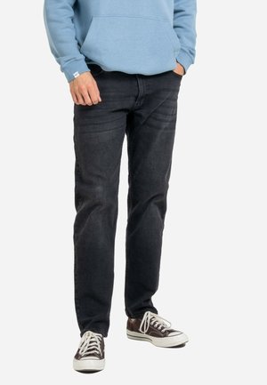 Homme portant un jean noir, des baskets marron et blanches, et un sweat à capuche bleu clair, une main dans la poche et l'autre le long du corps.