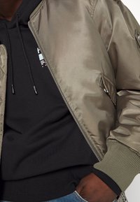 Olijfgroene bomberjack van glad satijn met rits, geribbelde manchetten en zijzakken. Zwarte hoodie eronder met aantrekkoorden.